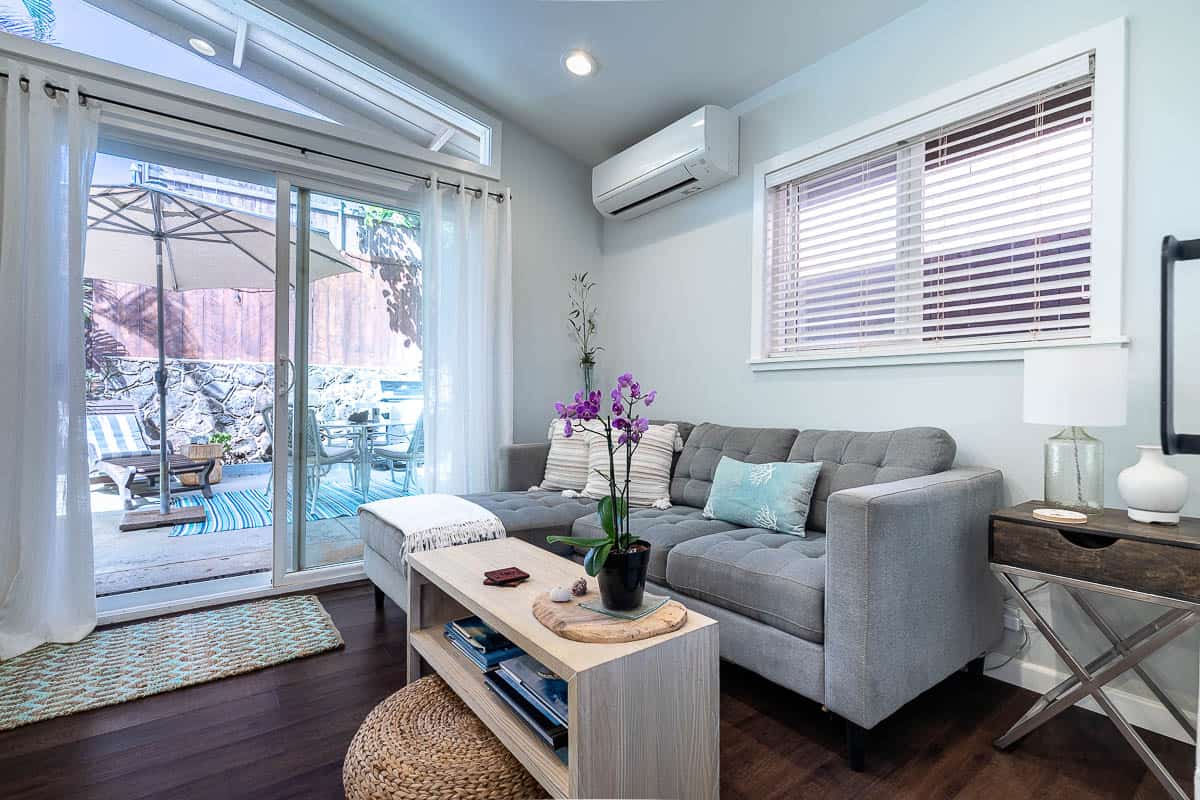 Living area with gray sectional sofa, coffee table, wall-mounted air conditioner, and sliding glass doors opening to a private patio.